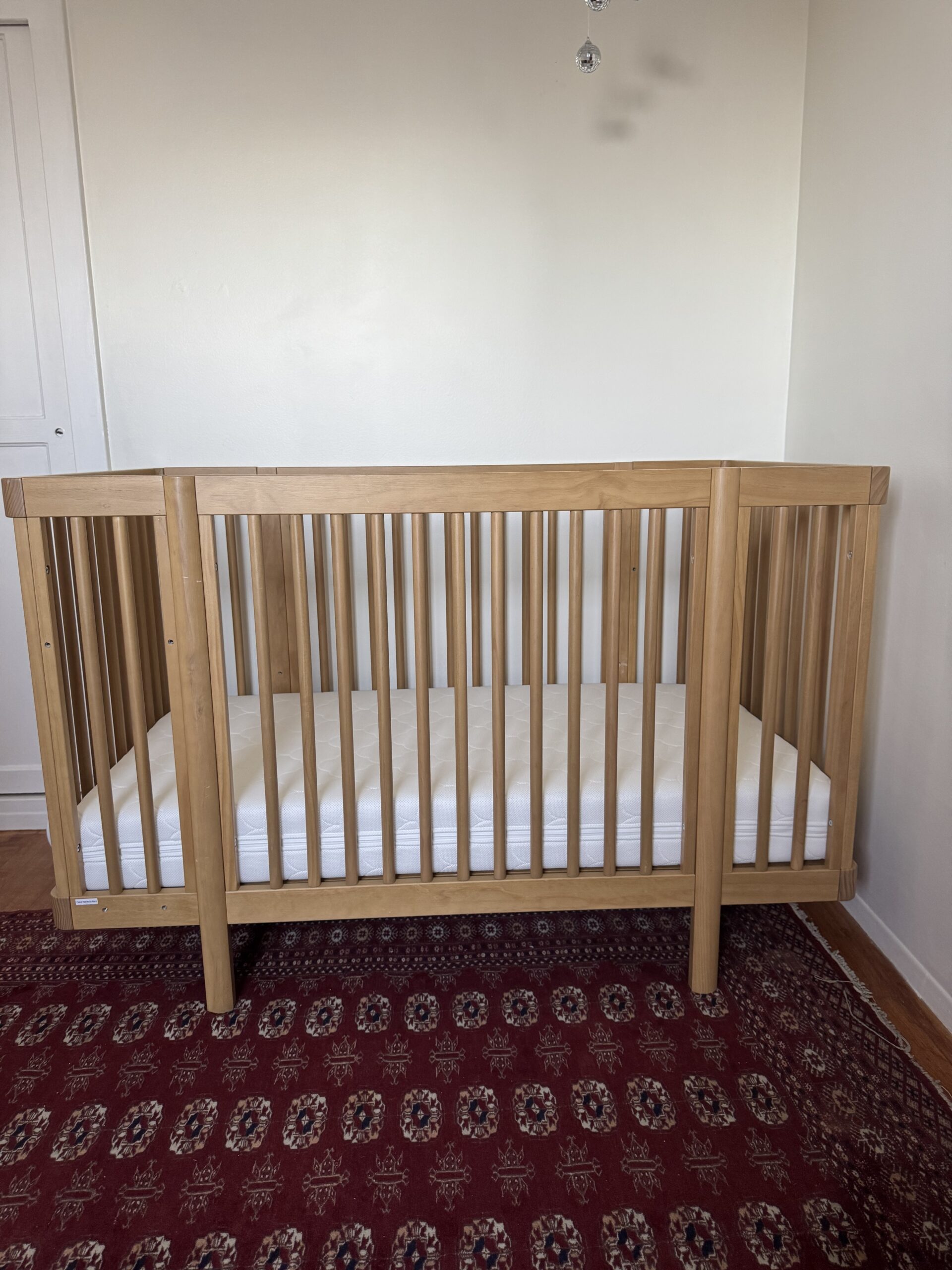 A wooden baby crib with vertical slats and a white mattress sits on a red patterned rug against a plain white wall.