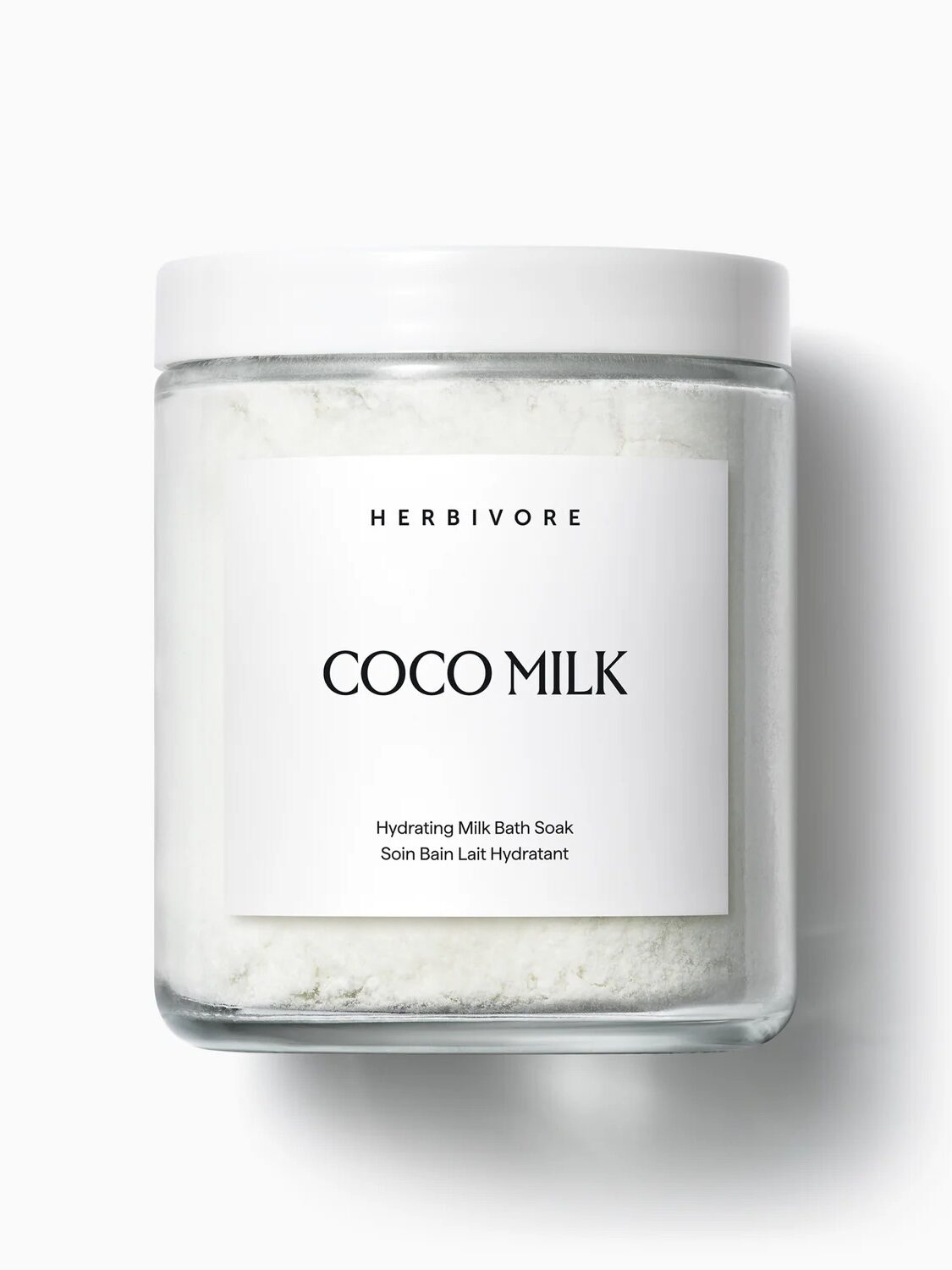 A clear glass jar with a white lid labeled "Herbivore Coco Milk Hydrating Milk Bath Soak" filled with white powder, shown against a plain white background.