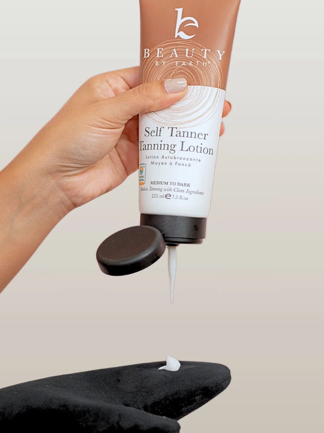 A hand squeezes self tanner lotion from a tube labeled "Beauty by Earth" onto a black glove.