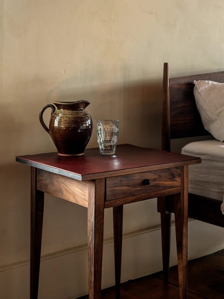 A brown ceramic pitcher and a clear glass sit on a wooden nightstand next to a bed with white bedding against a beige wall.