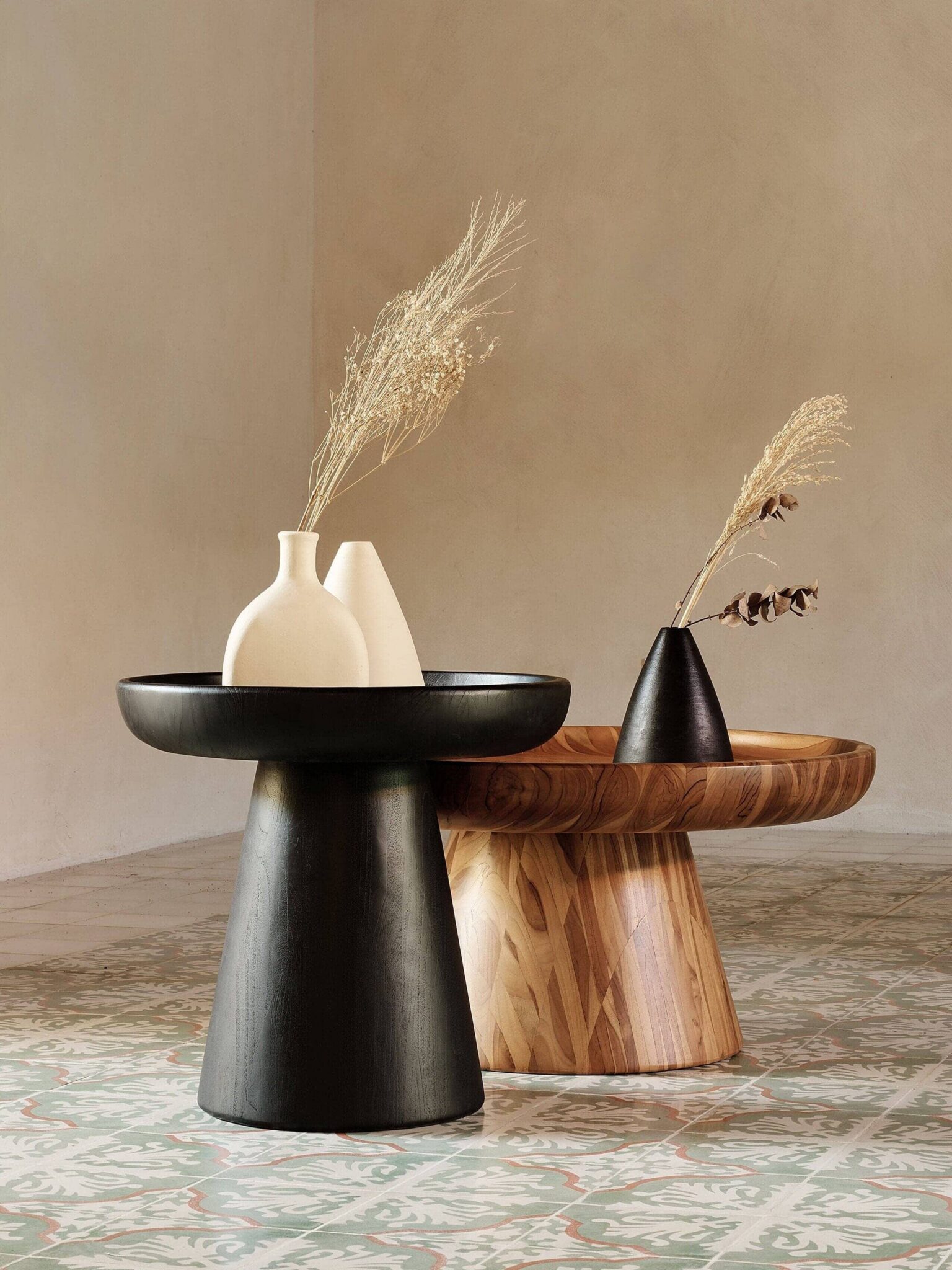 Two modern side tables, one black and one wood-toned, each holding minimalist vases with dried grasses, set on a patterned tile floor against a neutral wall.
