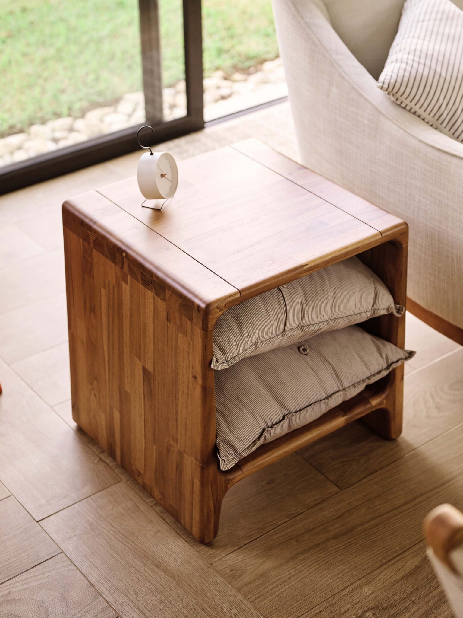 A wooden side table with two striped cushions stored inside and a small clock on top, placed next to a cream armchair by a large window.