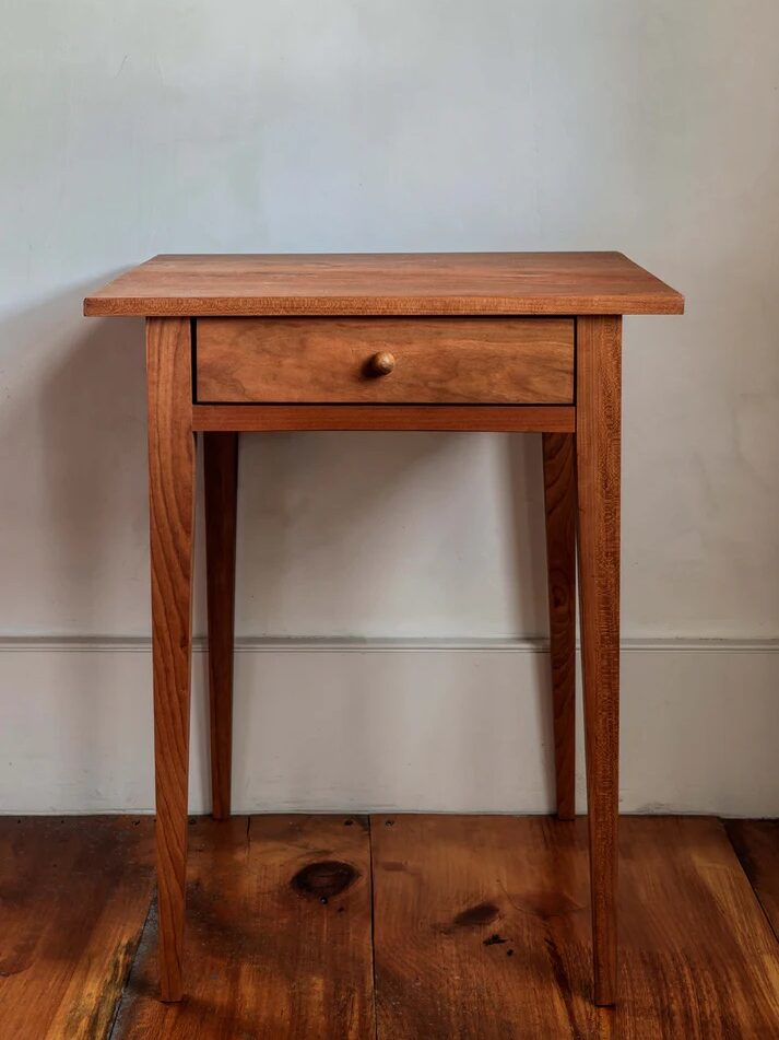 A simple wooden side table with a single drawer and four straight legs, placed on a hardwood floor against a plain light-colored wall.