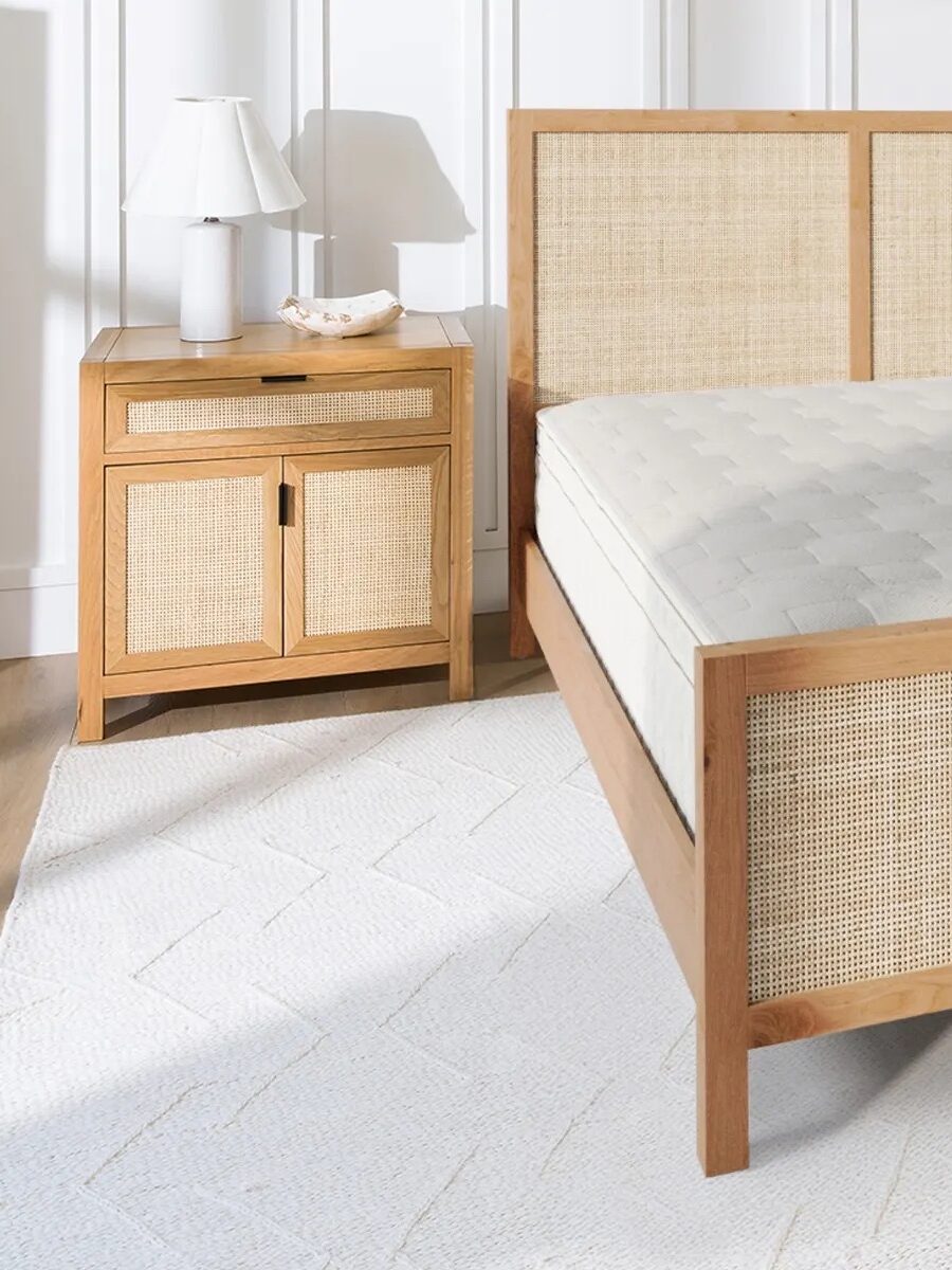 A wooden bed frame with a light woven headboard and footboard is next to a matching nightstand with a lamp, bowl, and white textured rug on the floor.