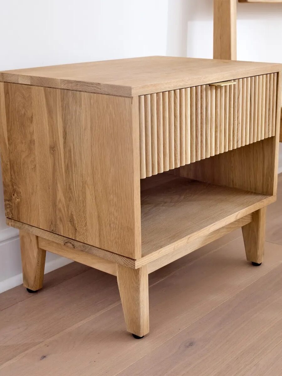 A wooden bedside table with tapered legs, a grooved drawer front, and an open shelf, placed on a light wood floor against a white wall.