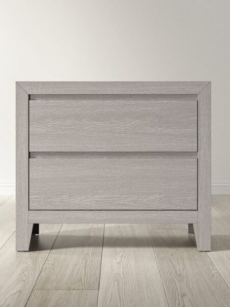 A light wood two-drawer nightstand sits on a light wooden floor against a plain white wall.