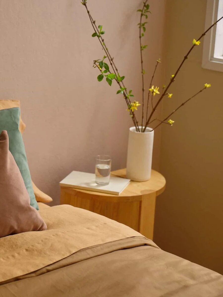 A neatly made bed with yellow, green, and brown pillows next to a round wooden nightstand holding a vase with branches, a book, and a glass of water.