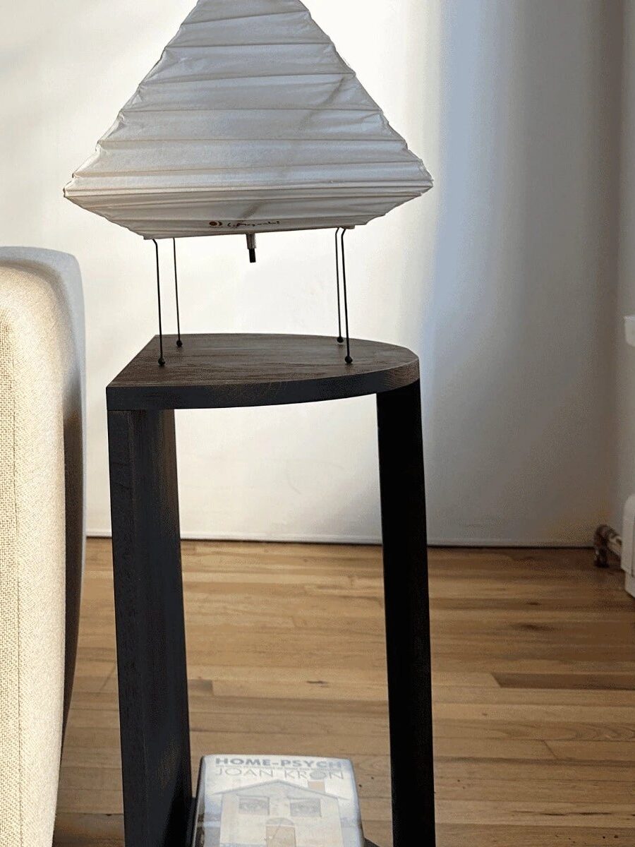 A beige sofa with brown and gray pillows sits next to a black side table holding a white geometric lamp; hardwood floor and a window are in the background.