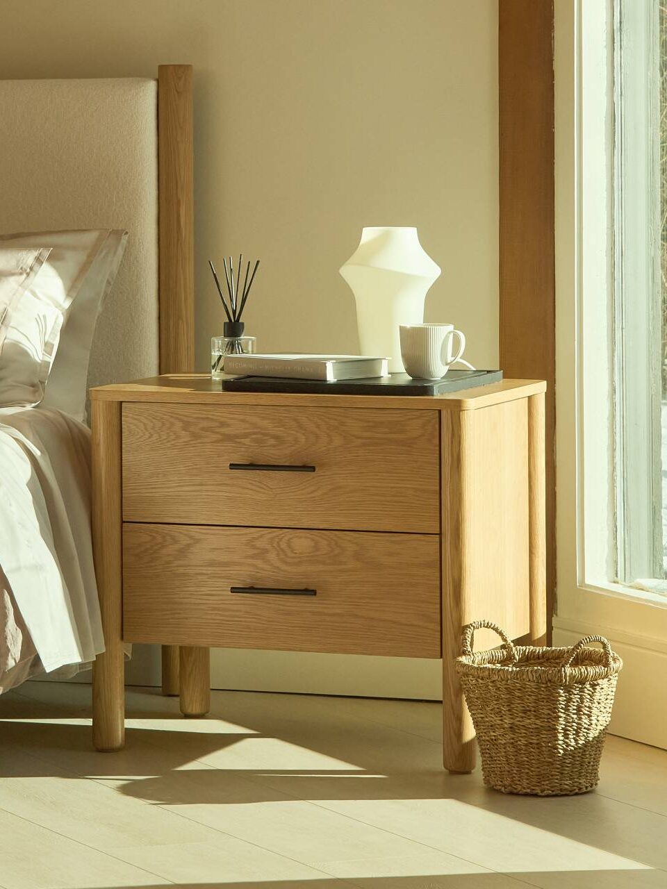 A wooden nightstand with two drawers sits beside a bed with beige bedding. On the nightstand are a white lamp, reed diffuser, book, and tray. A woven basket is on the floor nearby.