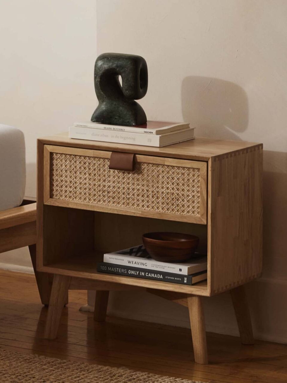 A wooden nightstand with angled legs, an open lower shelf, a drawer with a woven front and leather pull, topped with books, a small sculpture, and a bowl, beside a bed.
