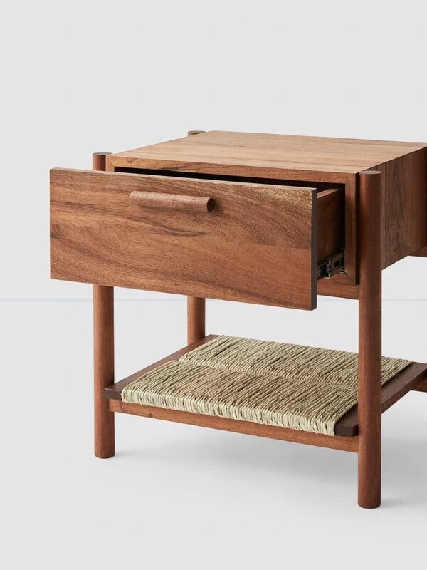 A wooden nightstand with one open drawer and a woven shelf underneath, set against a plain light background.