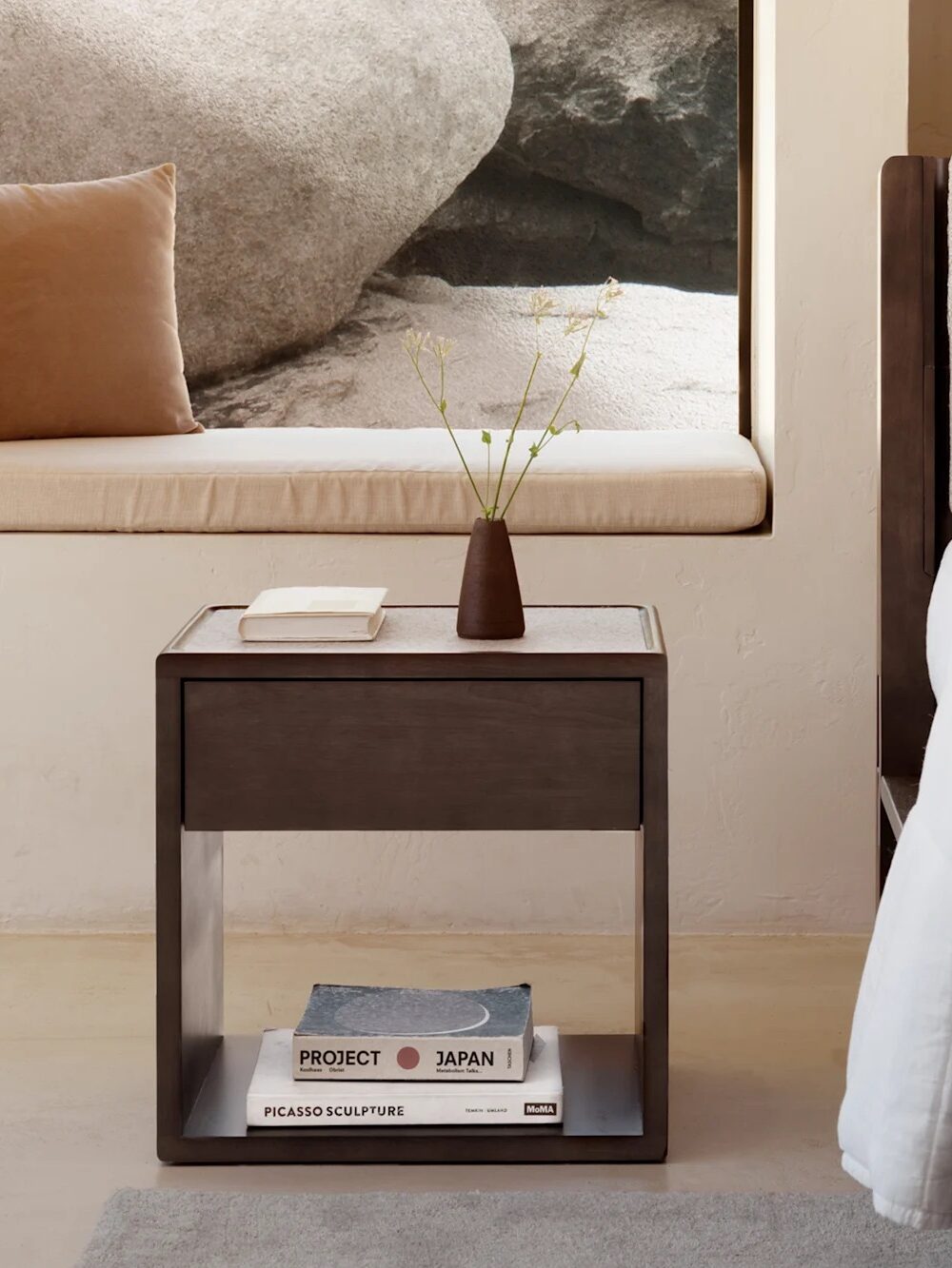 A modern bedroom with a bed, a wooden nightstand holding a vase and books, and a window seat with a cushion overlooking large rocks outside.