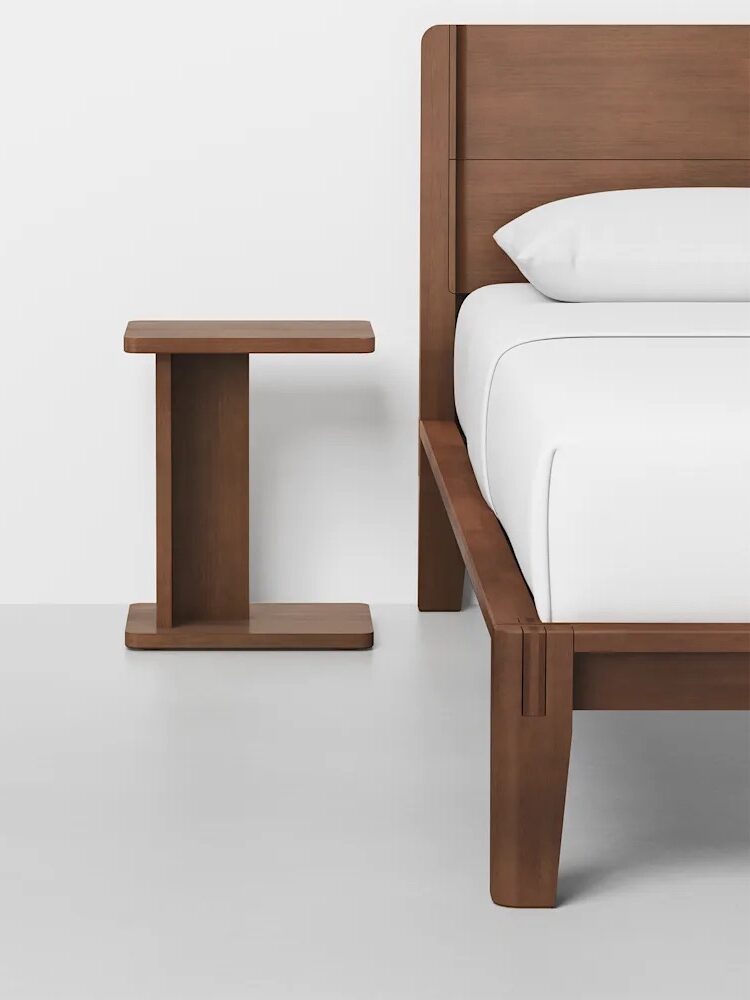 A wooden bed frame with white bedding next to a matching square wooden nightstand on a light gray floor against a white wall.