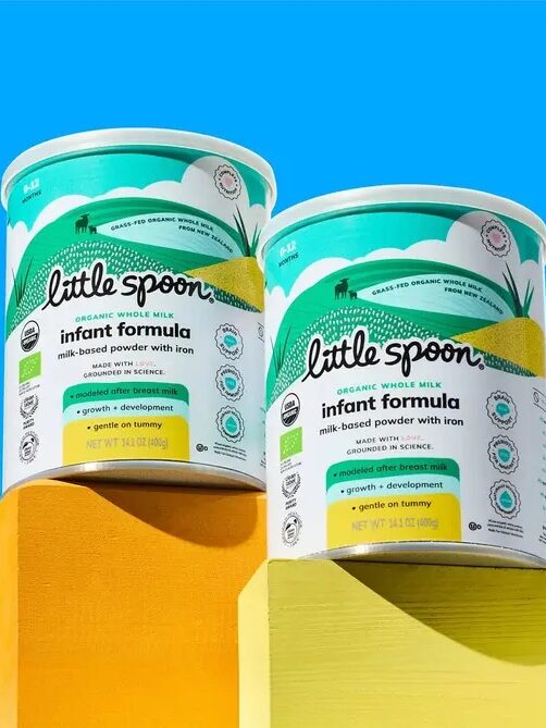 Two containers of Little Spoon organic whole milk infant formula sit on yellow blocks against a bright blue background.