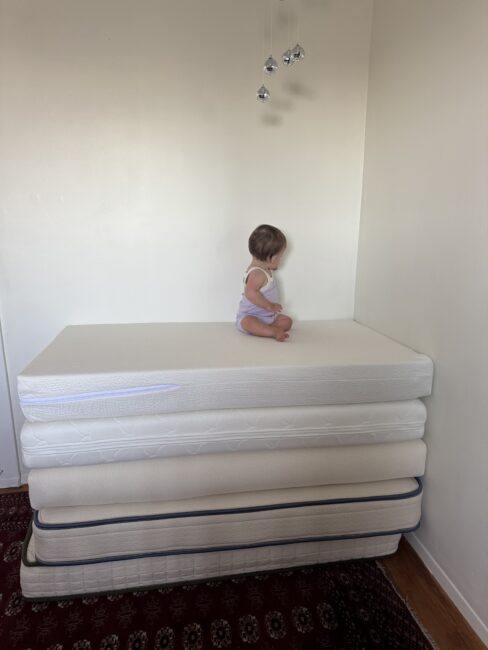 A small child sits on top of a stack of five mattresses in the corner of a sparsely furnished room with white walls.