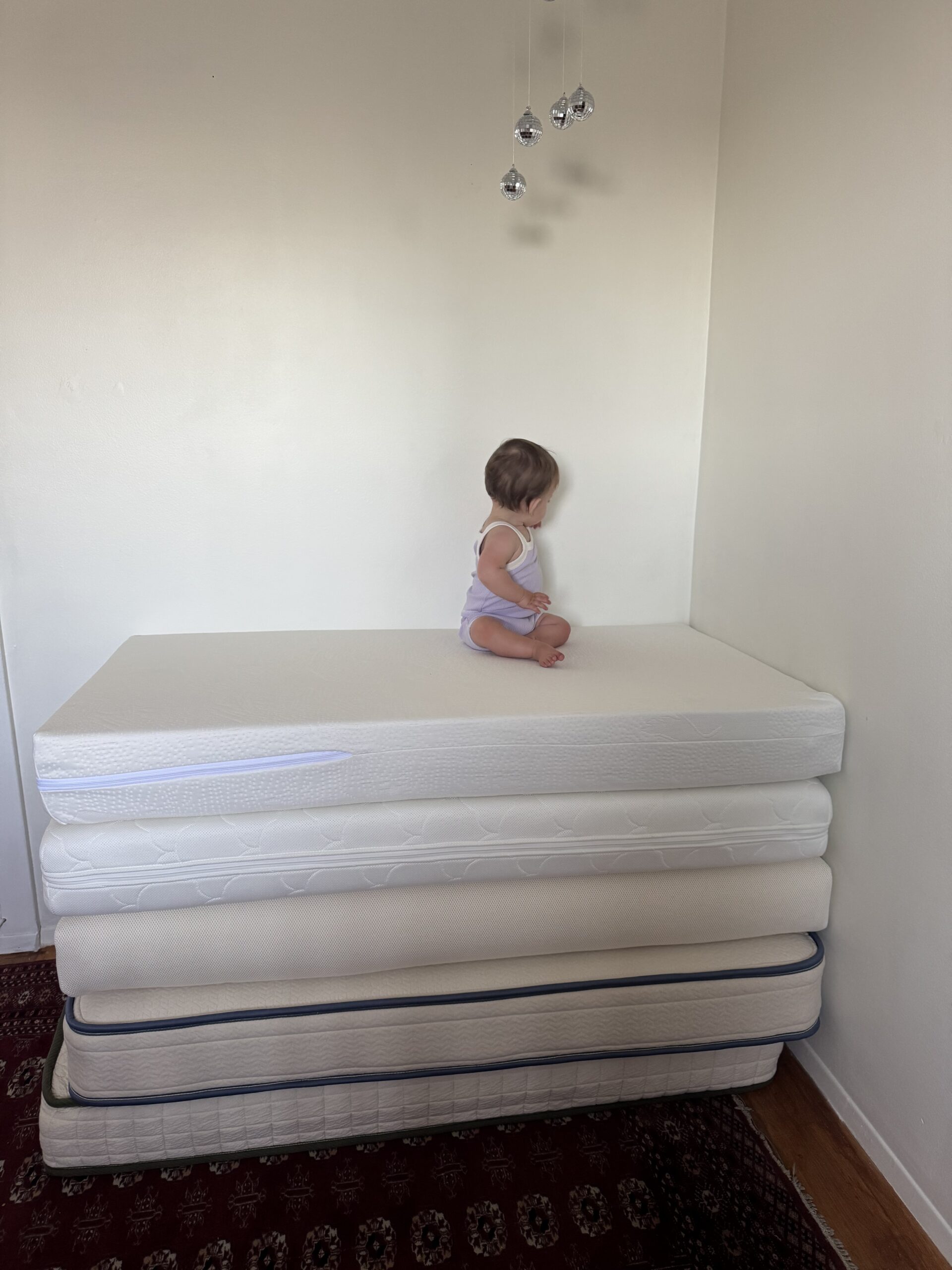 A small child sits on top of a stack of five mattresses in the corner of a sparsely furnished room with white walls.