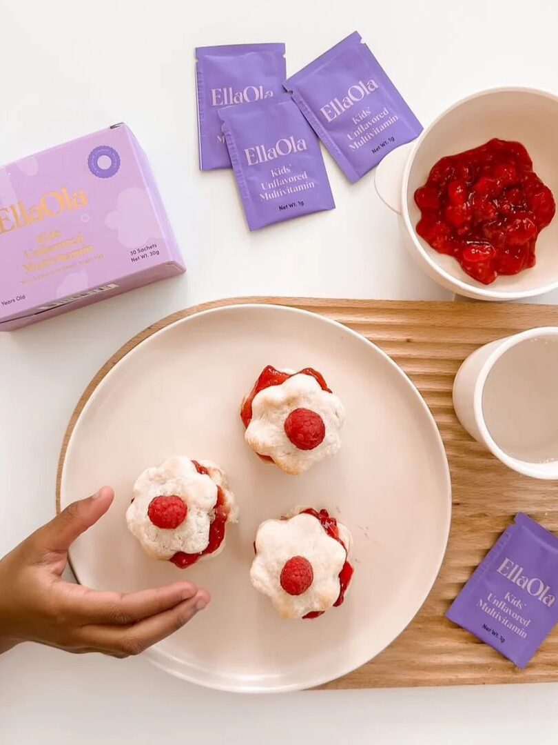 A hand reaches for a dessert topped with whipped cream and a berry; three desserts on a plate, a bowl of berry sauce, a cup, EllaOla sachets, and a box on the table.