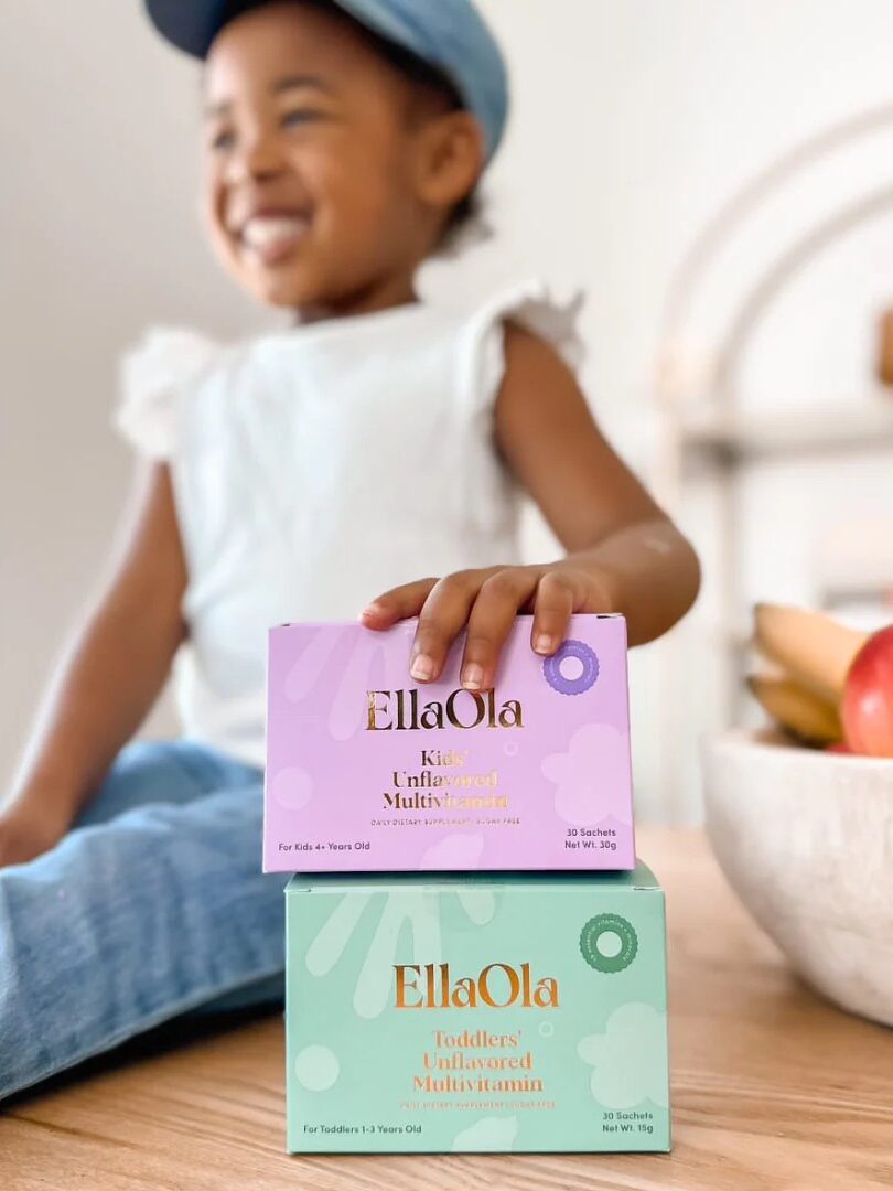 A smiling child sits at a table, holding up two boxes of EllaOla multivitamins, with a bowl of fruit in the background.