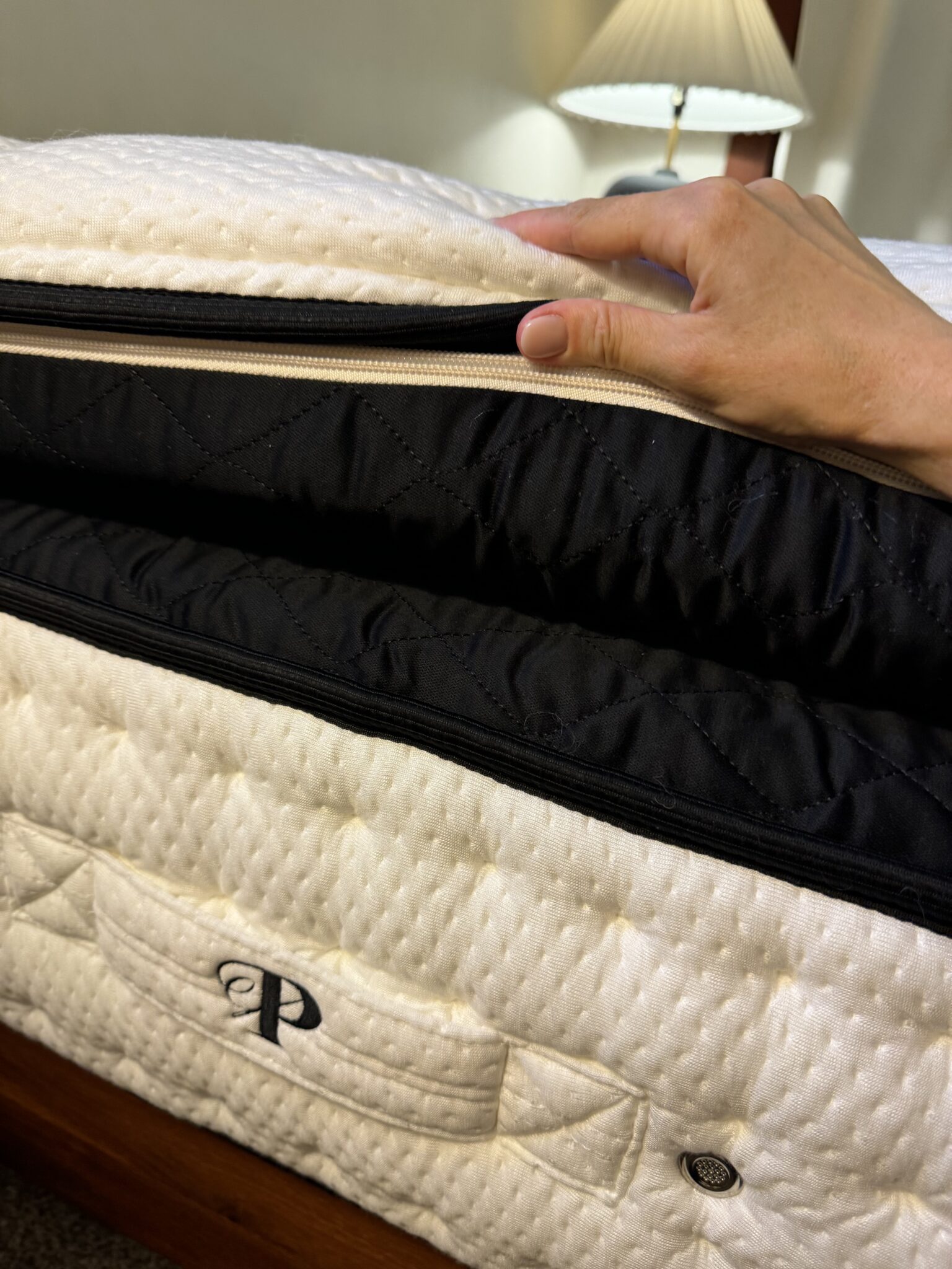 A hand opens the zipper of a latex mattress cover, revealing the inner layers. A quilted handle with the letter "P" is visible on the side.