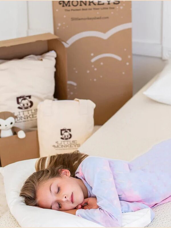 A young girl sleeps on a bed with a white pillow, while plush toys and branded packaging from Little Monkeys are visible in the background.