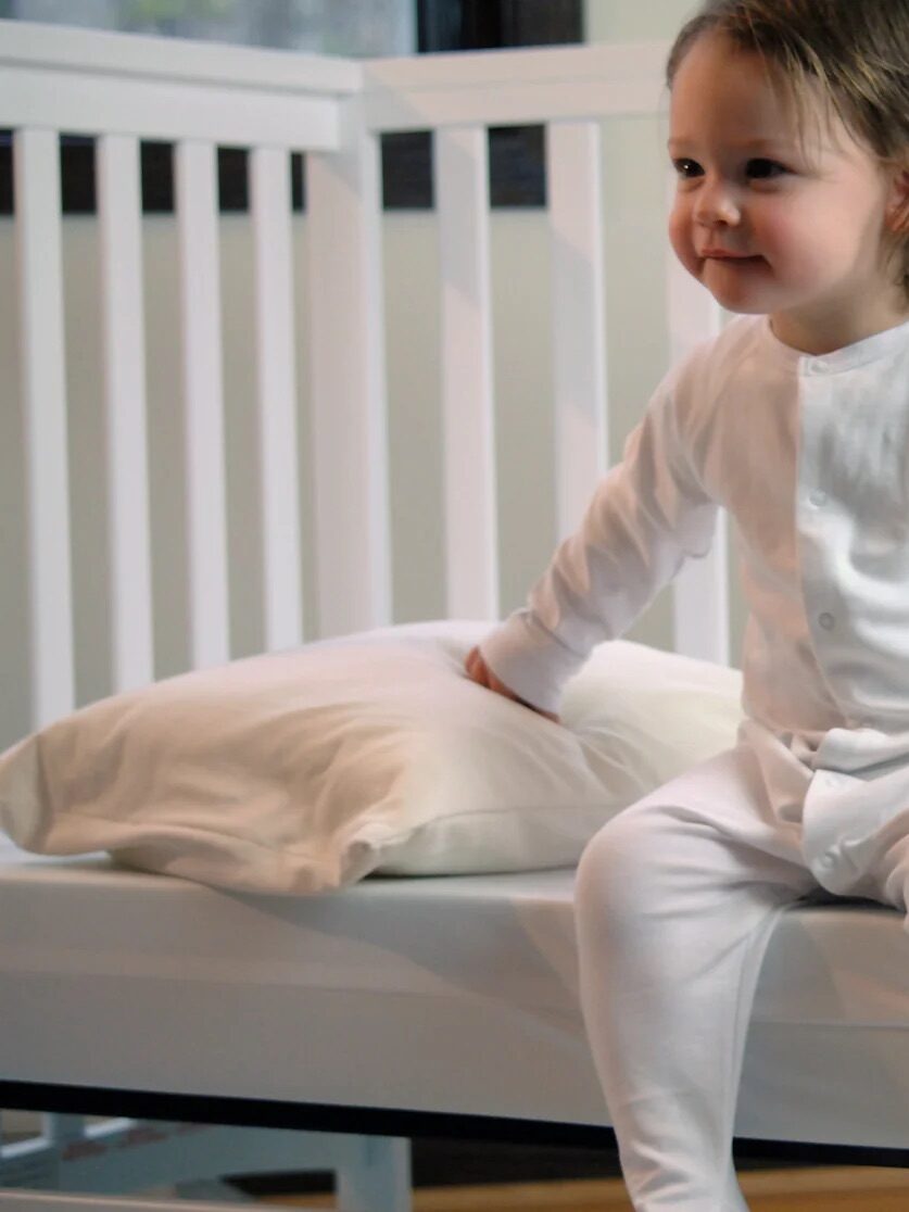 A young child in white pajamas sits on the edge of a bed with a white pillow, in front of a white crib.