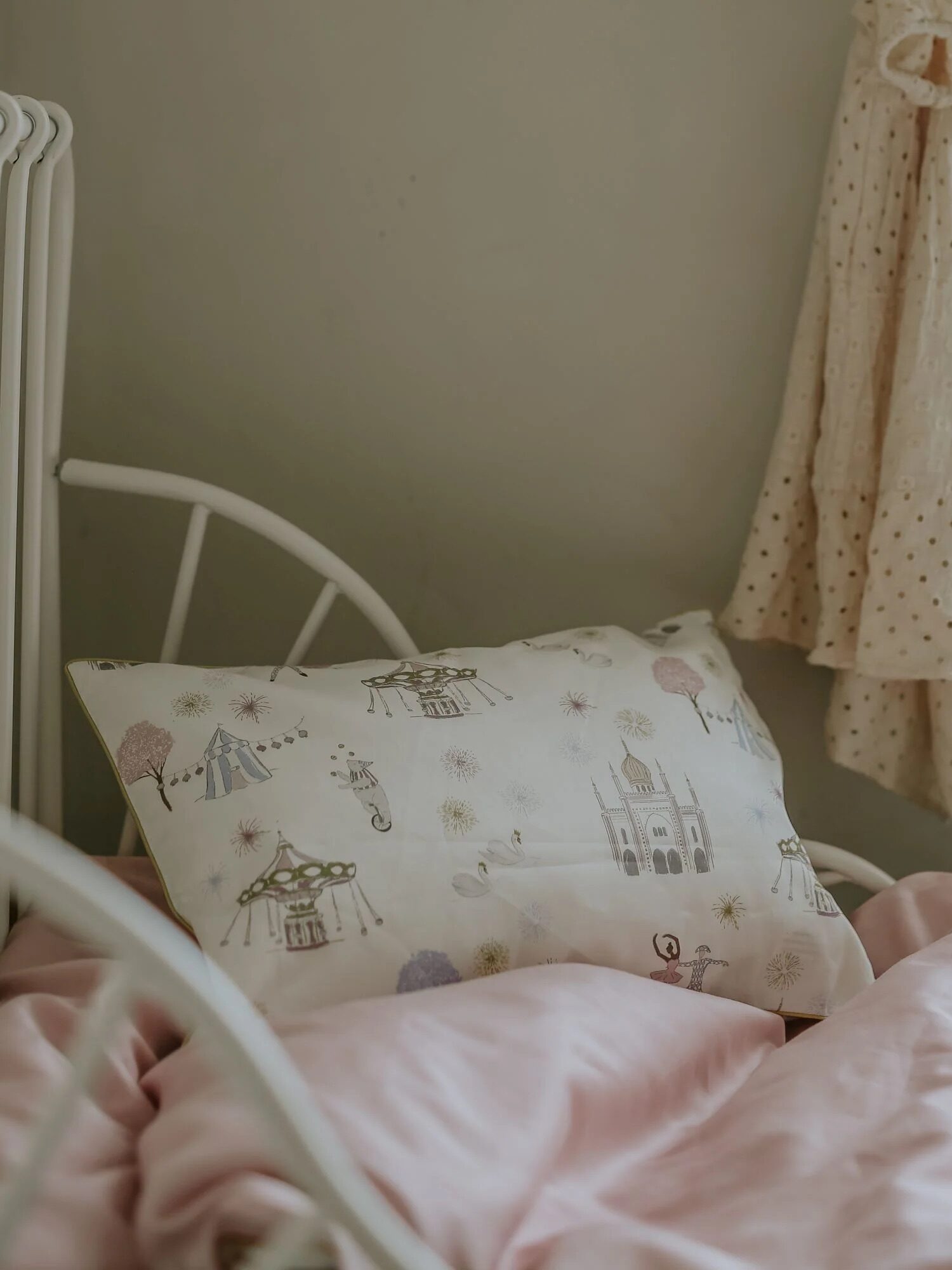 A child's white metal bed with a pink blanket, a printed pillow featuring whimsical designs, and a cream-colored dress hanging on the wall.