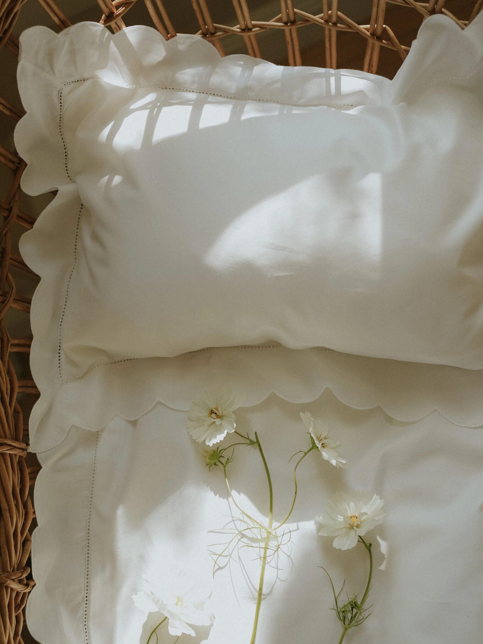 A white pillow with ruffled edges rests on a wicker chair, with white flowers and sunlight casting shadows across the fabric.