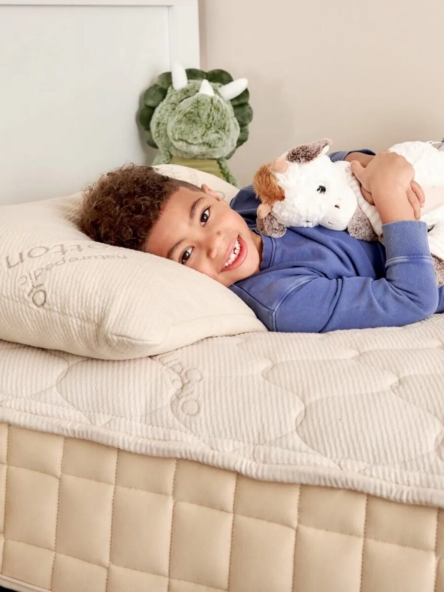 A child lies on a beige mattress, smiling while hugging a stuffed animal. The bed has a cream pillow and a quilted cover, with a green plush toy in the background.