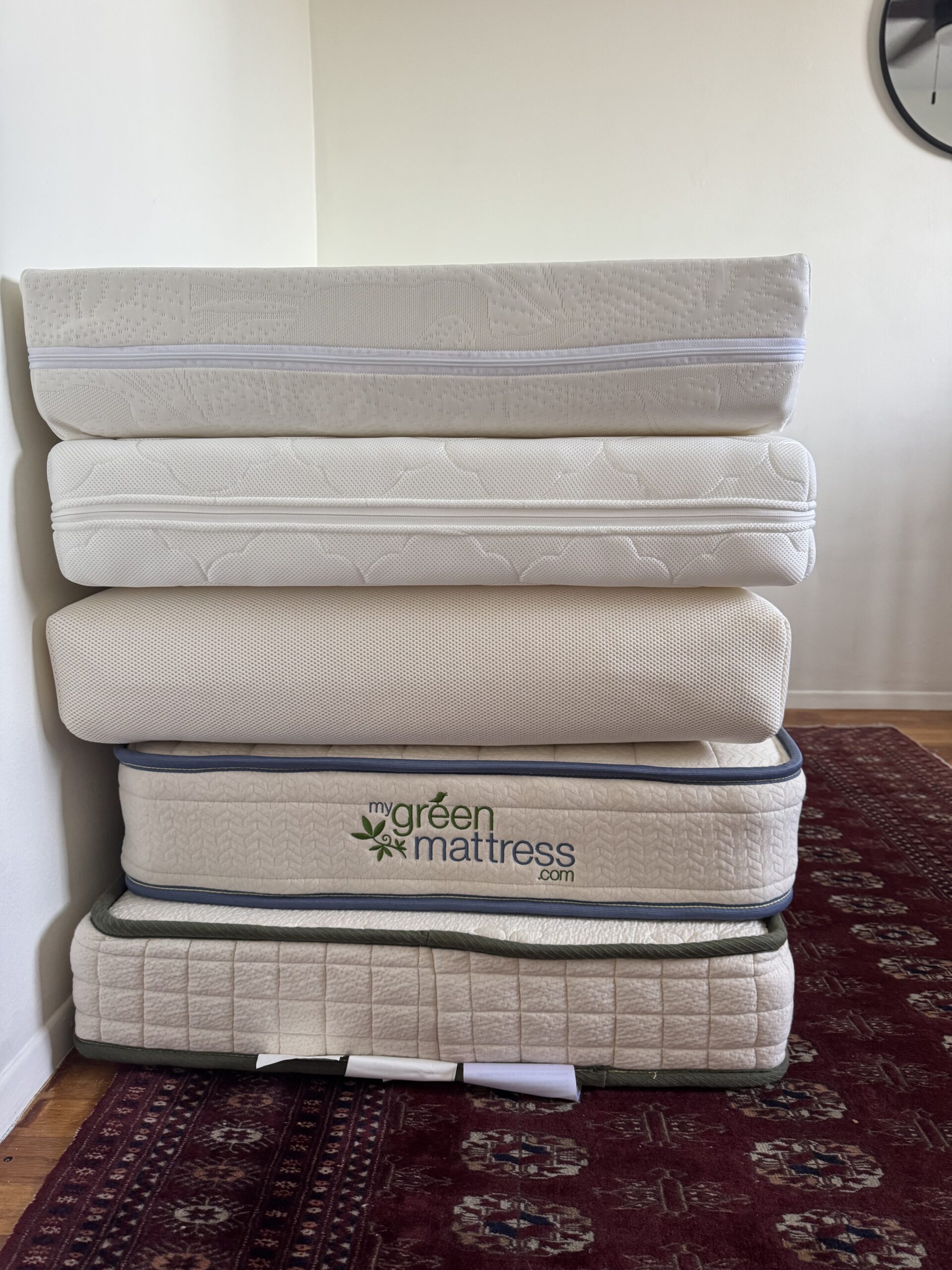 Five mattresses stacked against a white wall on a red patterned rug, with the "my green mattress.com" logo visible on one mattress.