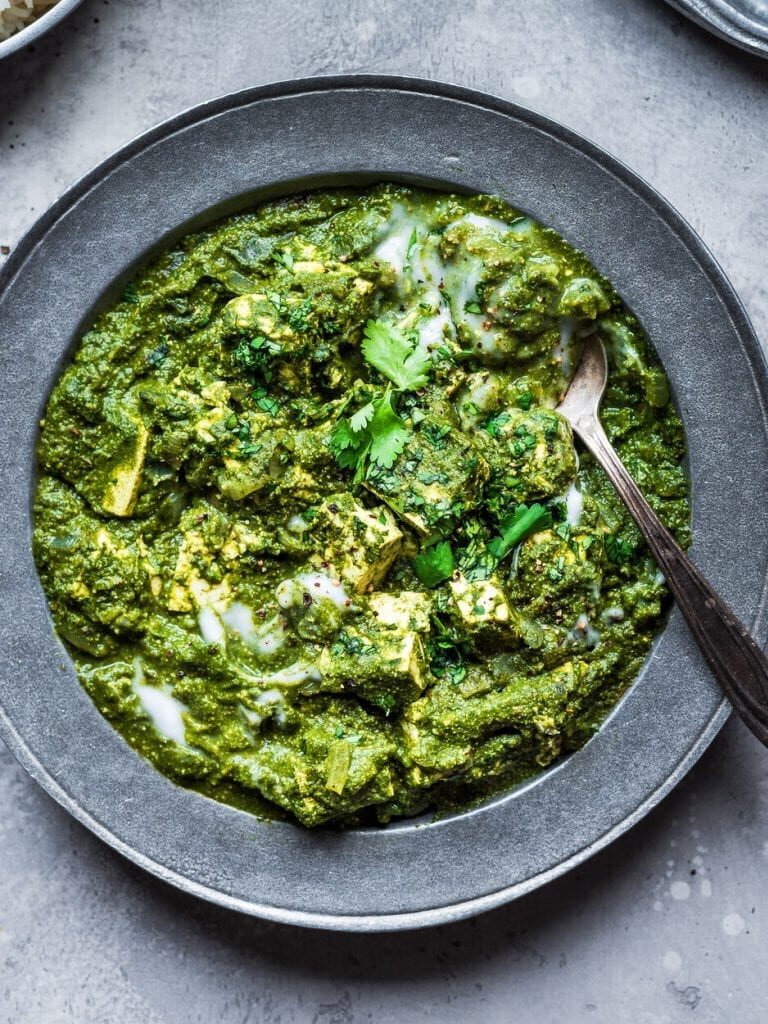 A bowl of palak paneer, an Indian spinach and cheese curry, garnished with cilantro and a drizzle of cream, with a spoon in the bowl.