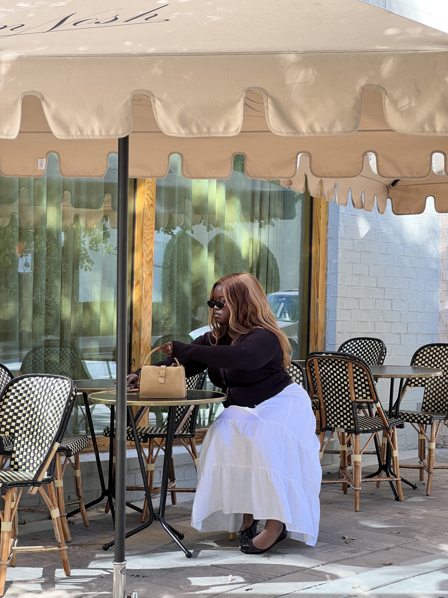 A woman in a black top and white skirt sits alone at an outdoor café table, reaching into her tan handbag under a beige umbrella.