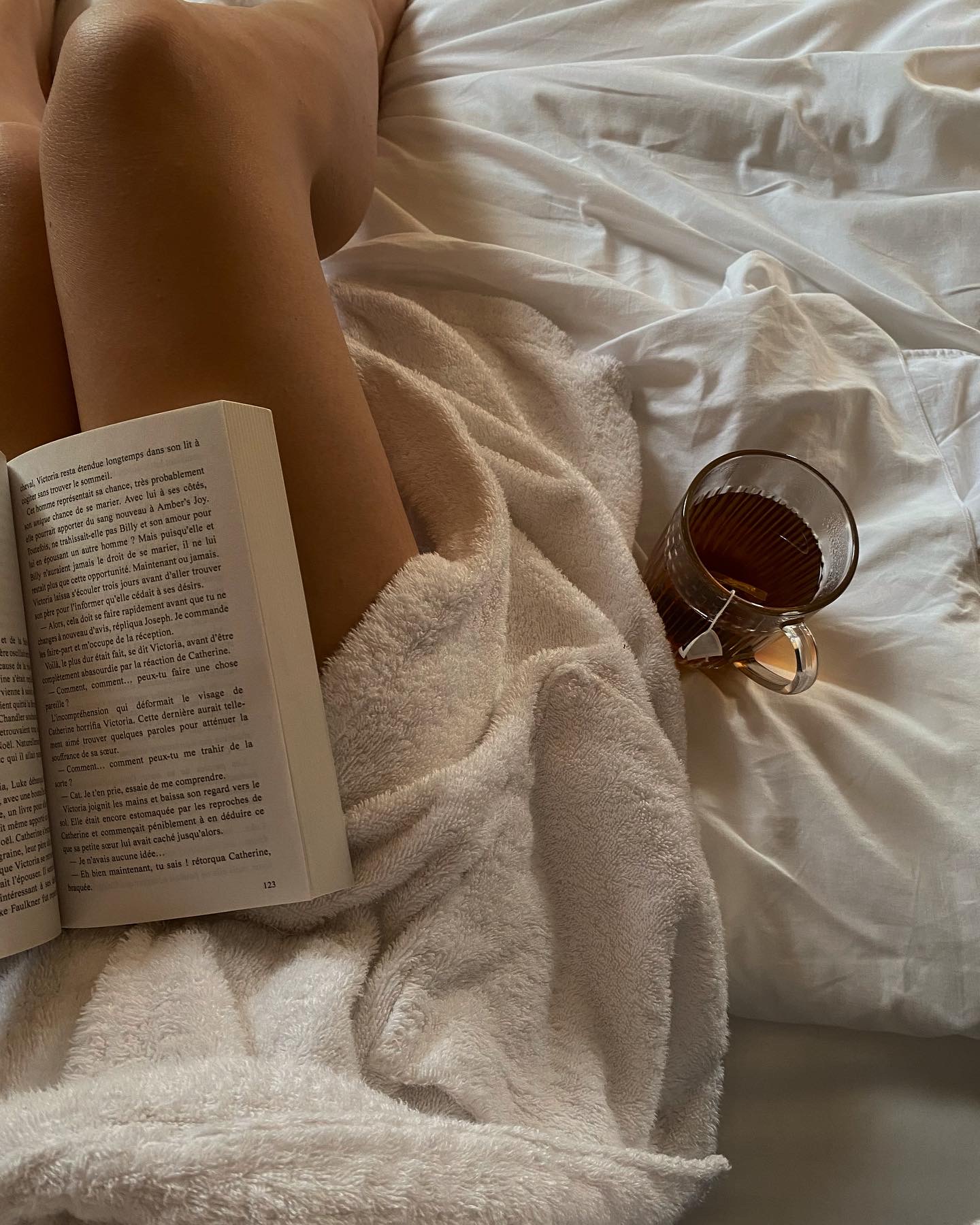 Person sitting on a bed covered with a light towel, reading a book in French, with a glass cup of tea placed on white sheets beside them.