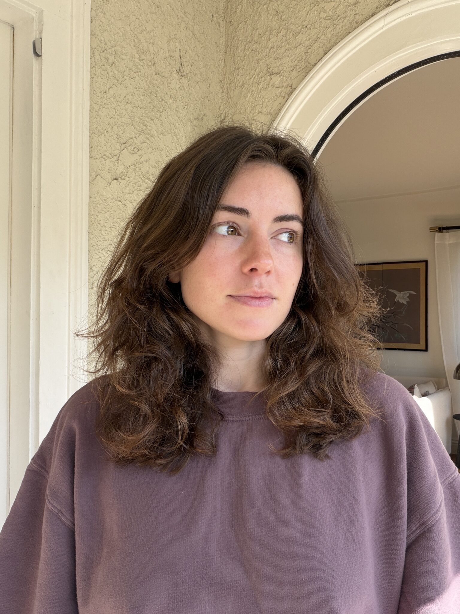 A woman with wavy brown hair wearing a mauve sweatshirt stands indoors, looking to her left near an arched doorway.