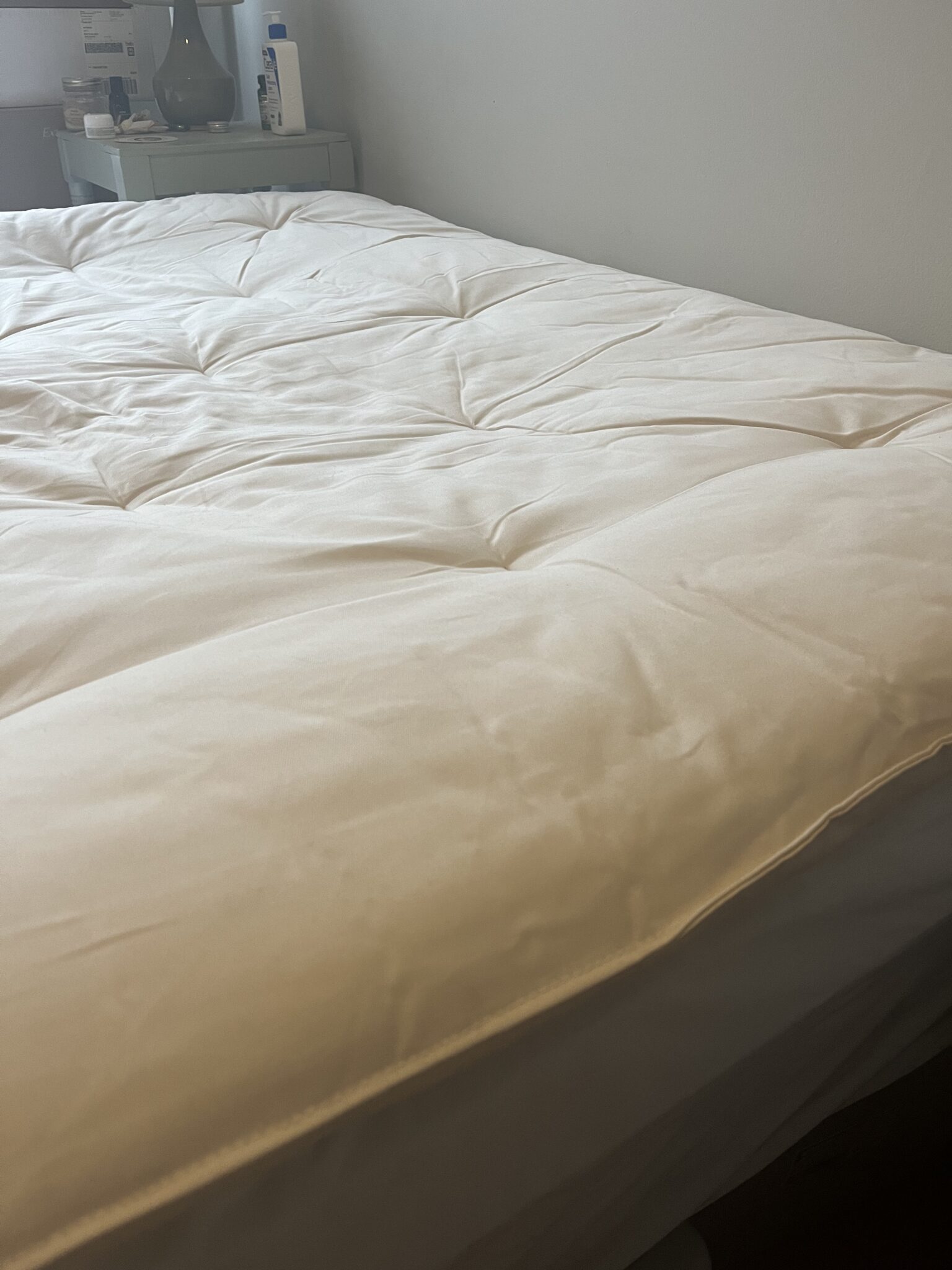 A neatly made bed with a white comforter next to a nightstand holding a green lamp, lotion bottle, and other small items.