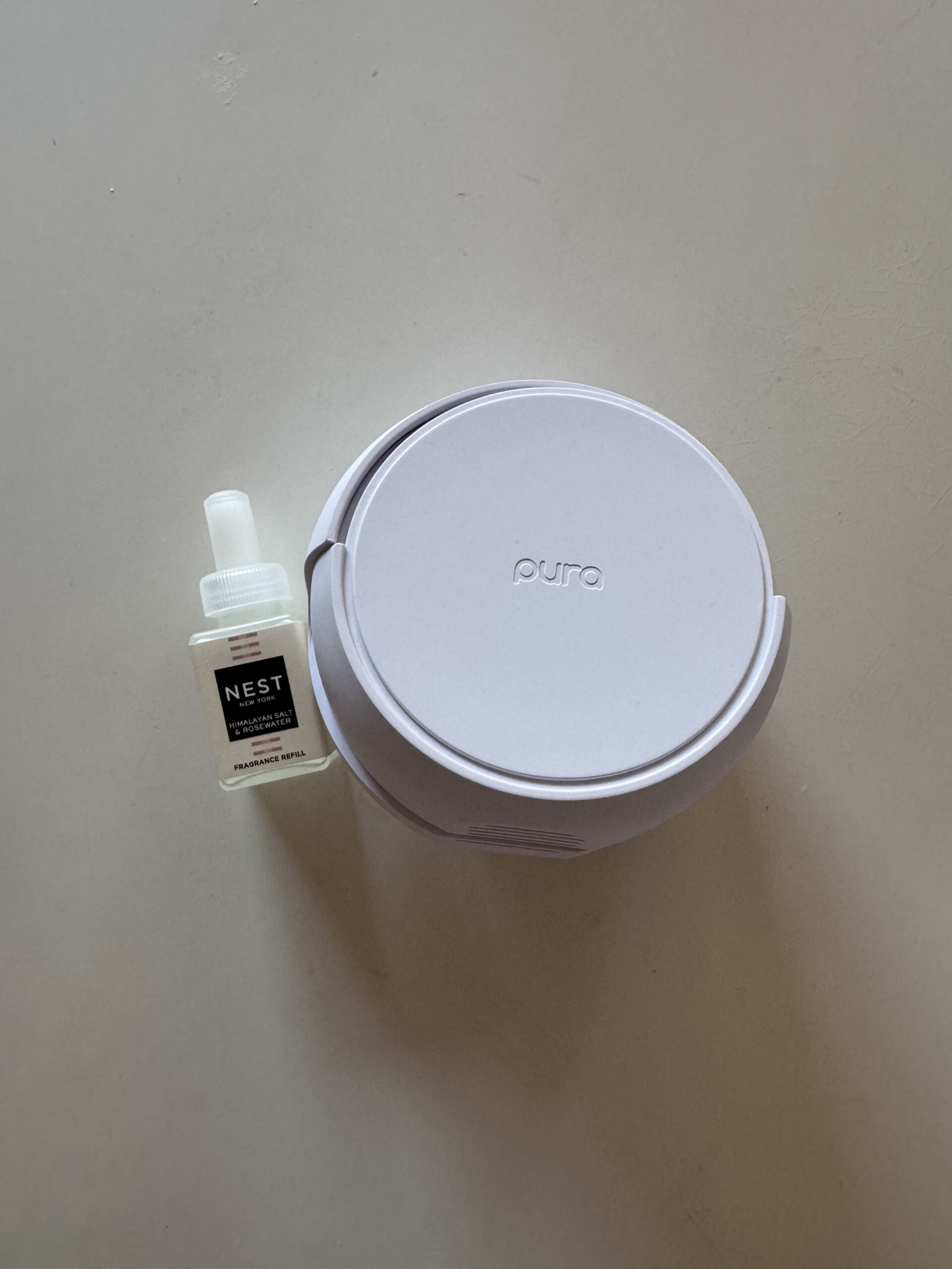 A white Pura smart home fragrance diffuser sits next to a small bottle of Nest New York Grapefruit fragrance refill on a light surface.