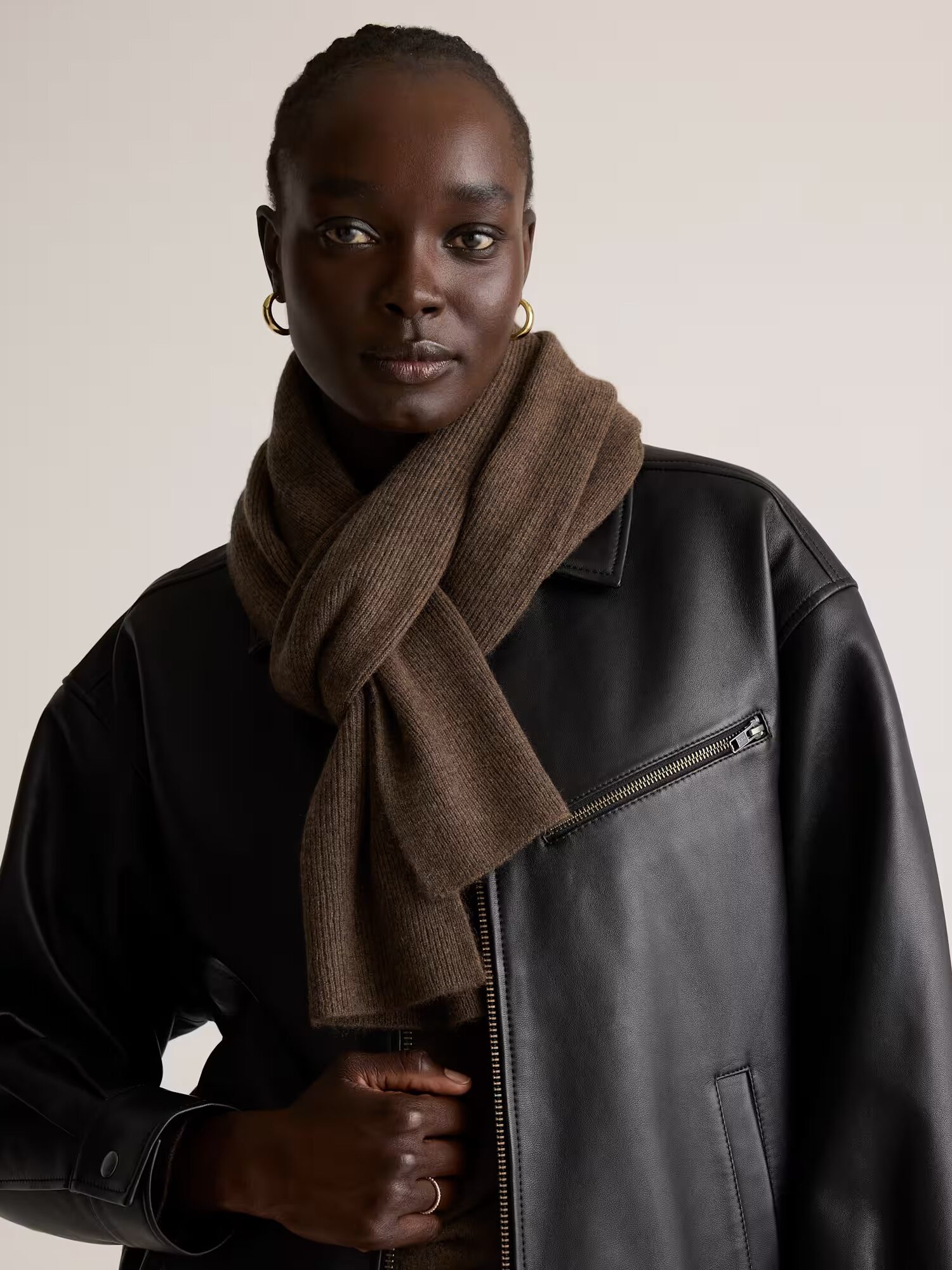 A person wearing a black leather jacket and a brown knit scarf, standing against a plain beige background.