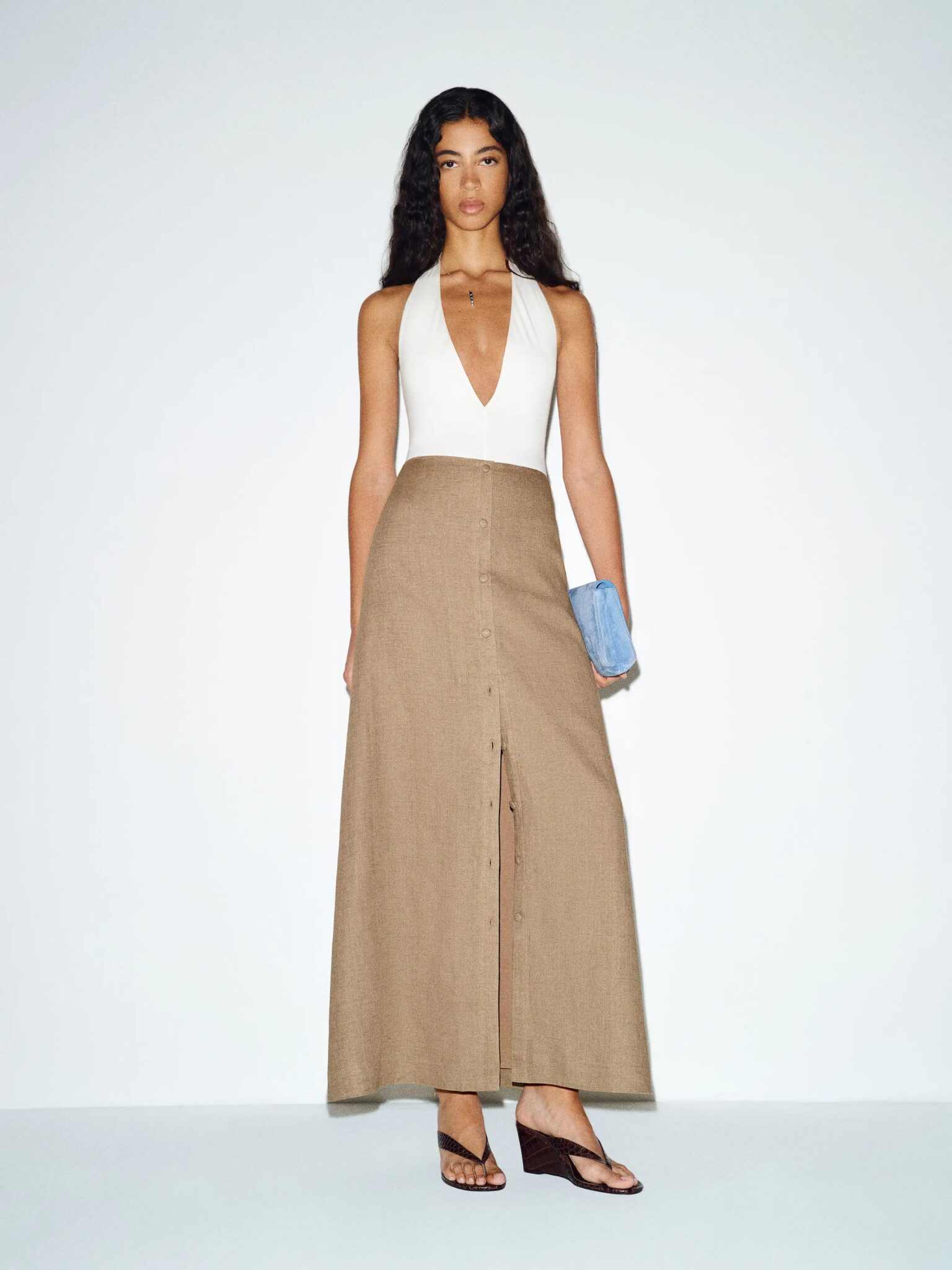 A woman stands against a plain background wearing a sleeveless white halter top and a long beige buttoned skirt, holding a blue clutch and wearing black sandals.