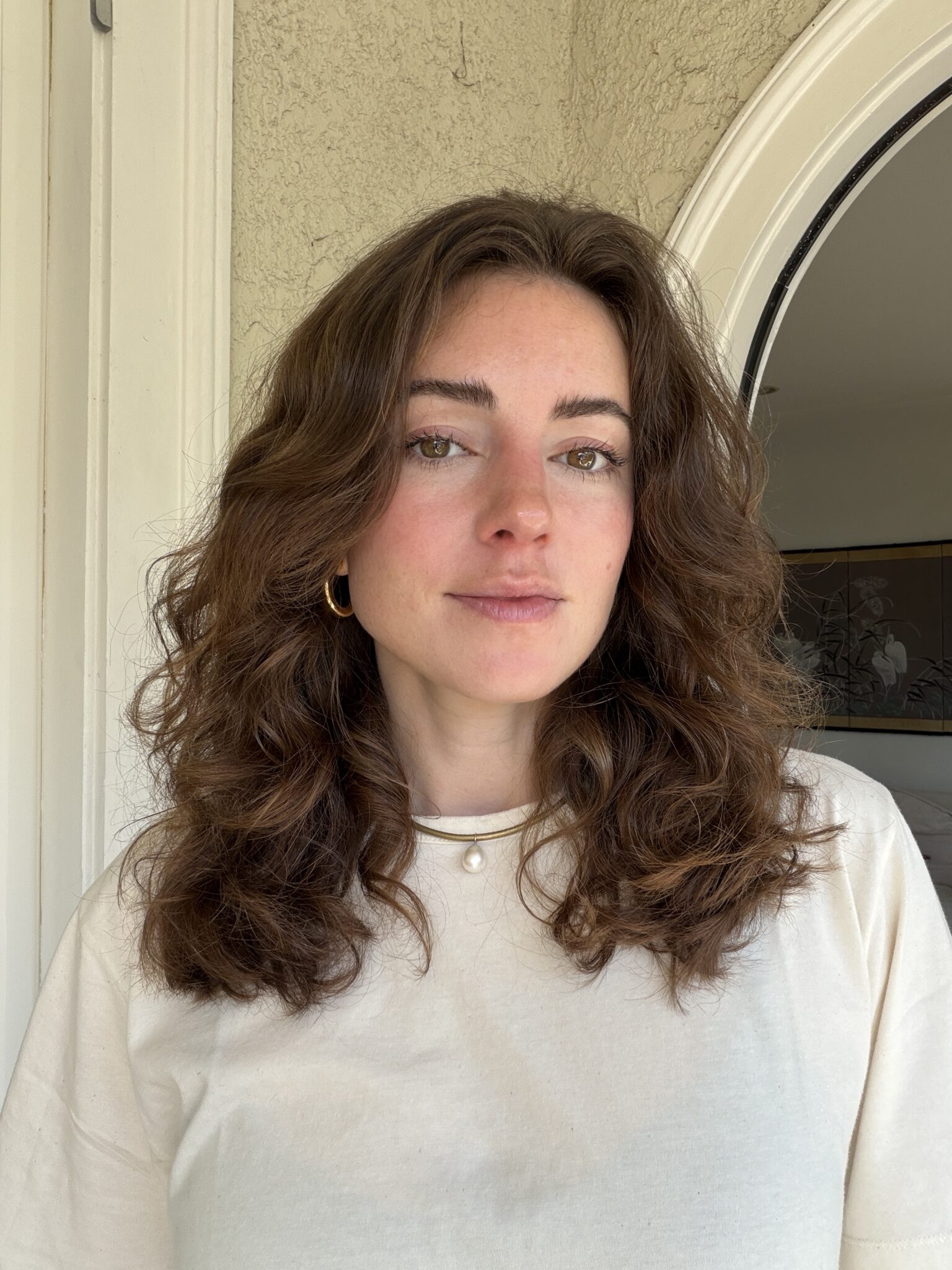 A woman with wavy brown hair wearing a white shirt and gold hoop earrings stands indoors in front of a light-colored wall and an arched doorway.