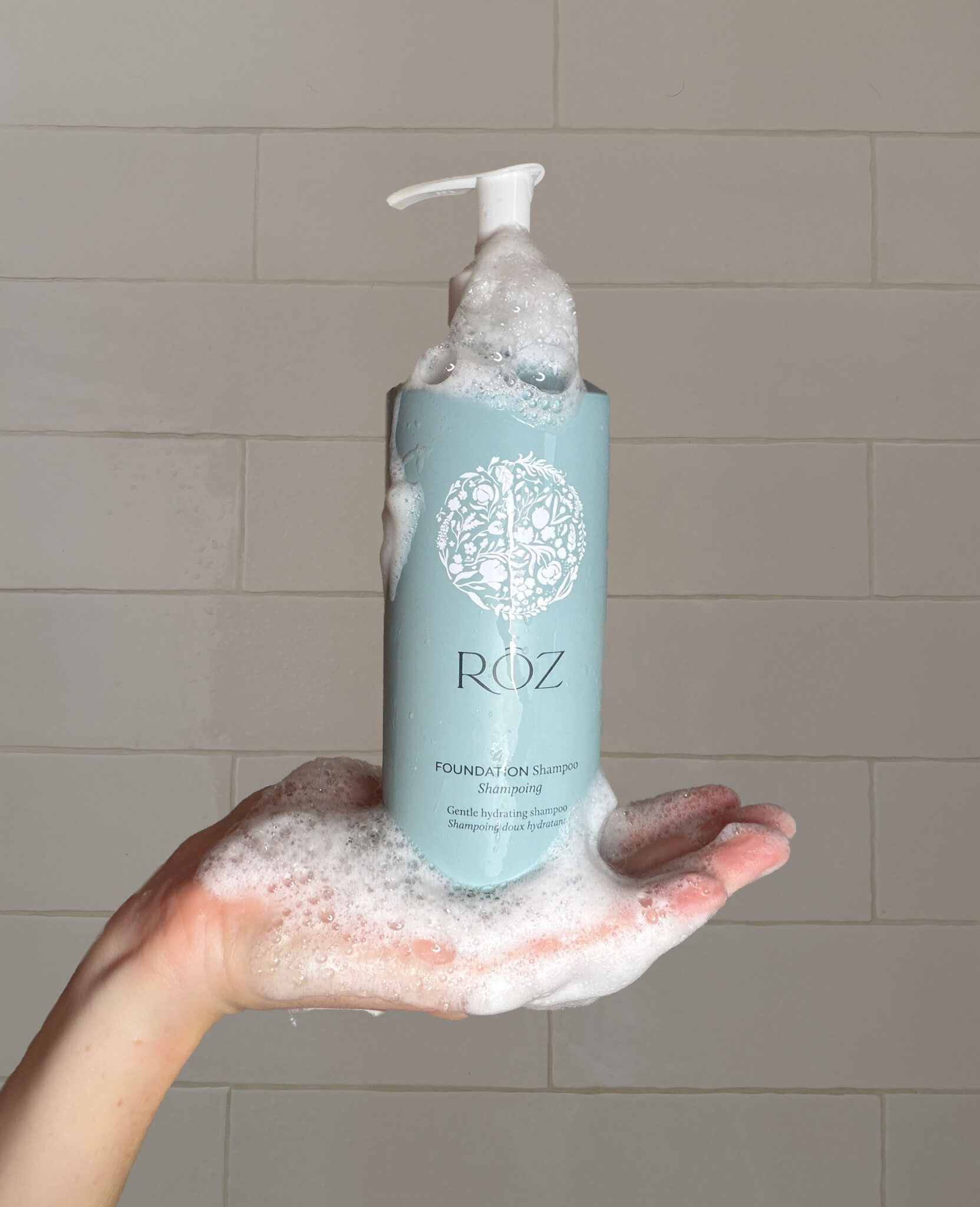 A hand holds a blue bottle of ROZ Foundation Shampoo, covered in foamy soap suds, against a tiled wall background.