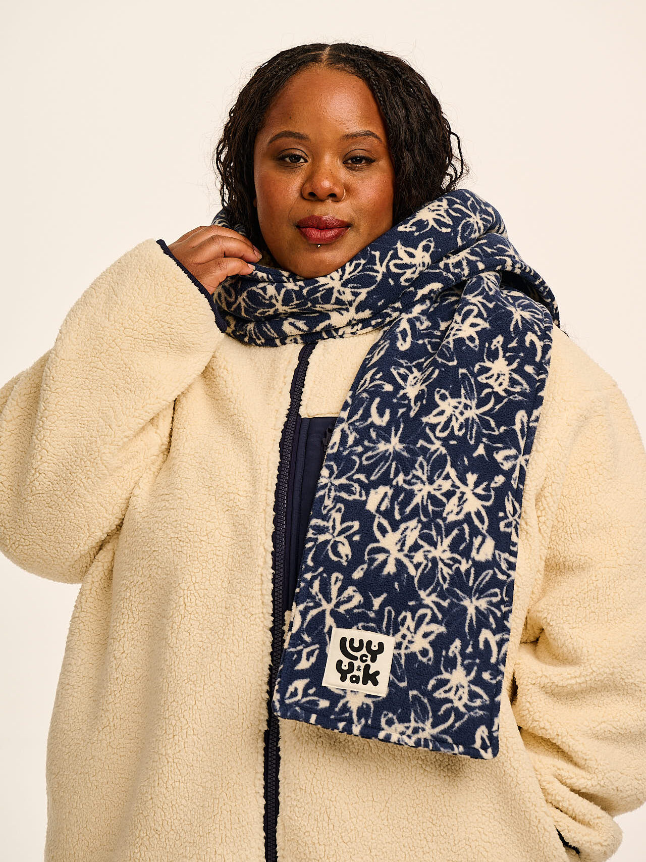 A woman wearing a cream sherpa jacket and a blue scarf with a white floral pattern stands against a plain background.