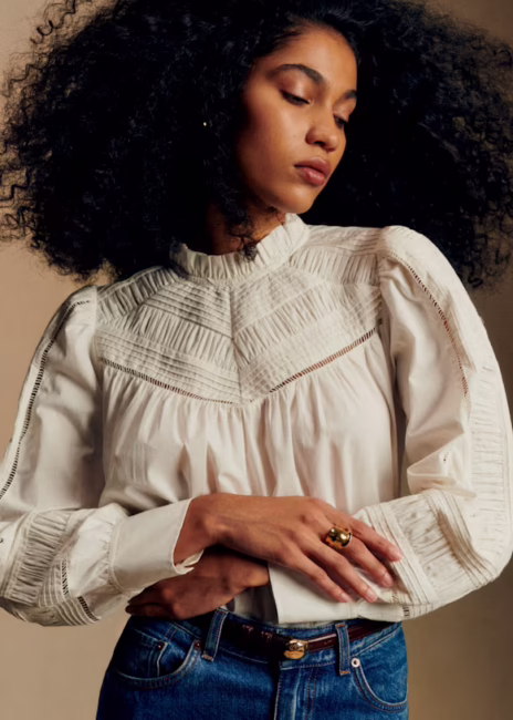 Woman with curly hair wears a cream-colored, long-sleeved blouse with intricate detailing and blue jeans, standing with arms crossed and looking down.