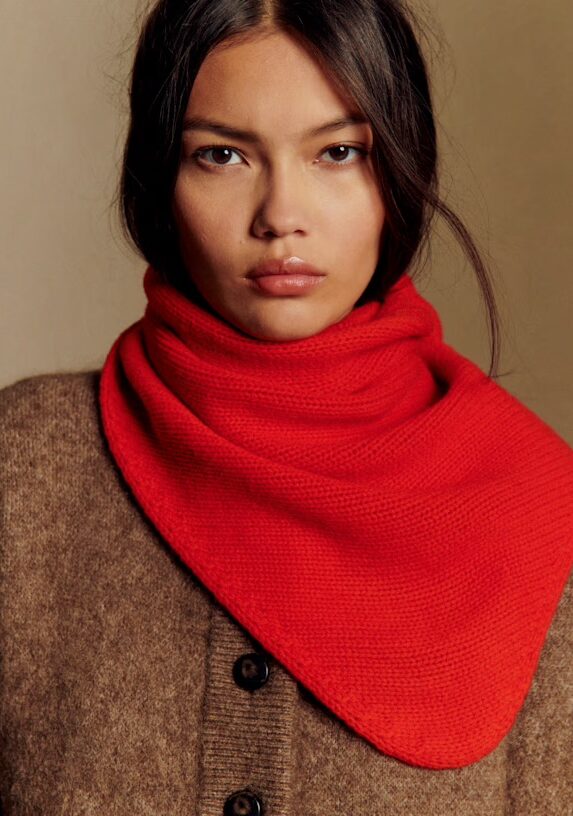 A woman with long dark hair wears a brown sweater and a bright red knitted scarf around her neck, looking directly at the camera with a neutral expression.
