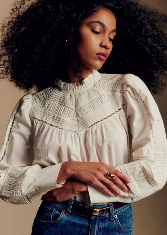 Woman with curly hair wears a cream-colored, long-sleeved blouse with intricate detailing and blue jeans, standing with arms crossed and looking down.