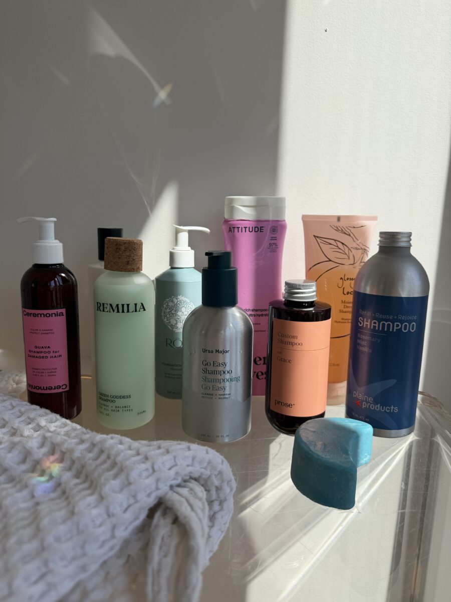 Various hair care products in colorful bottles and bars arranged on a clear surface with a folded white towel, lit by sunlight from the right.