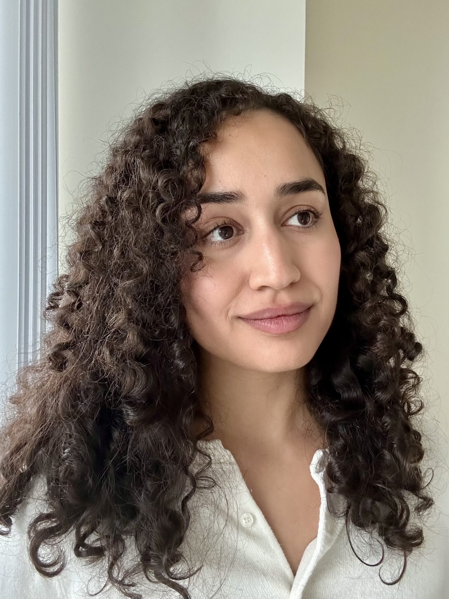 A woman with curly dark hair and a white shirt looks slightly to the side, standing indoors near a window with soft natural light.