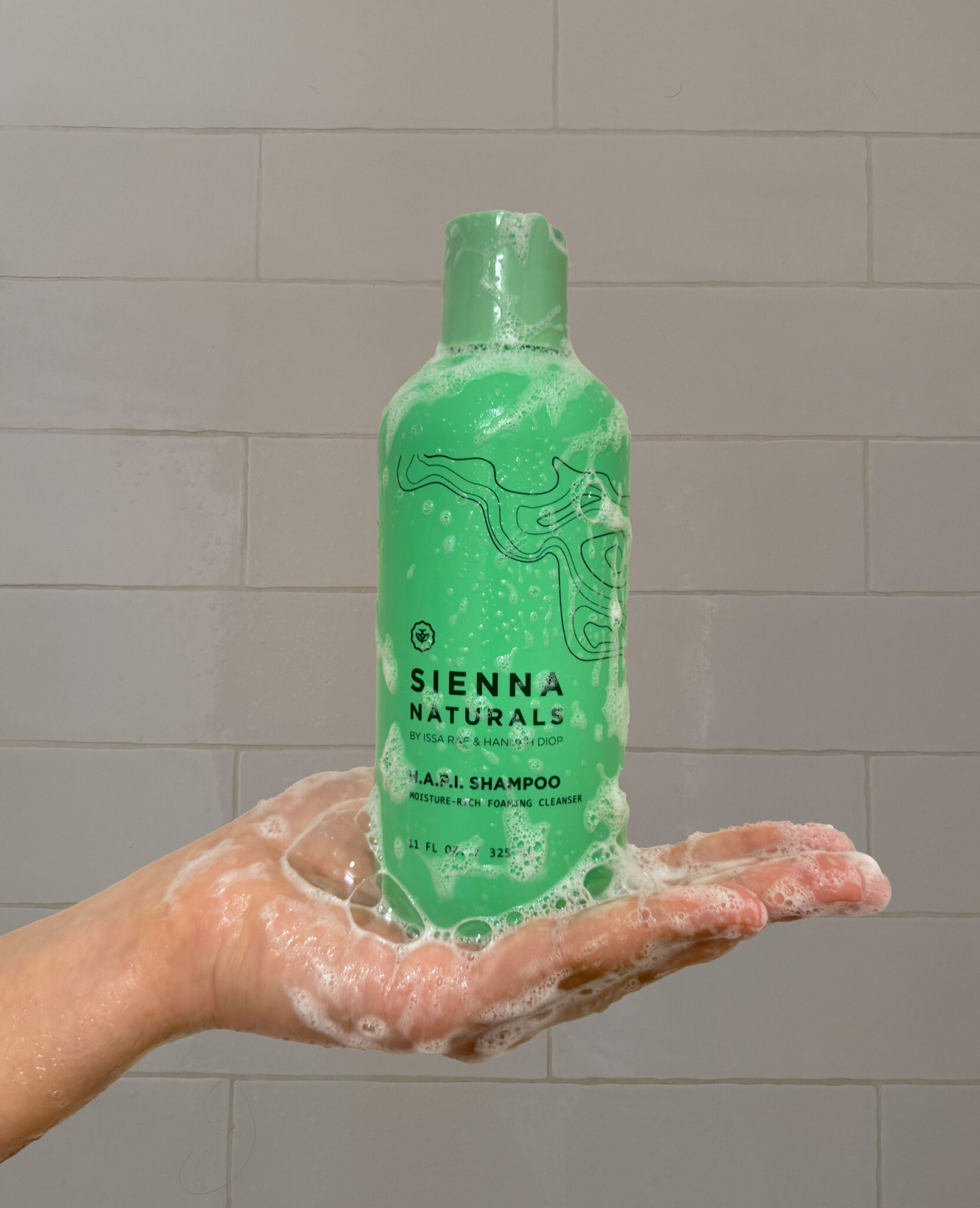 A hand covered in suds holds a green bottle of Sienna Naturals H.A.P.I. Shampoo in front of a tiled shower wall.