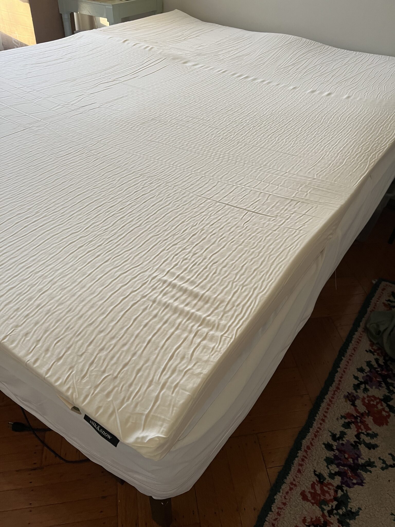 A bed with a white fitted sheet is split in the middle, revealing two separate adjustable bases; the bed is on a wooden floor next to a patterned rug.