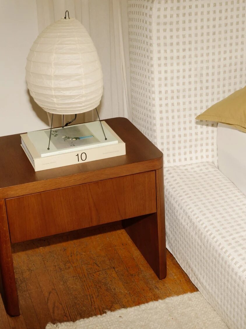 A wooden nightstand with a paper lantern lamp, books, and a pen sits beside a bed with beige pillows and a patterned headboard. The floor is wood with a light rug.