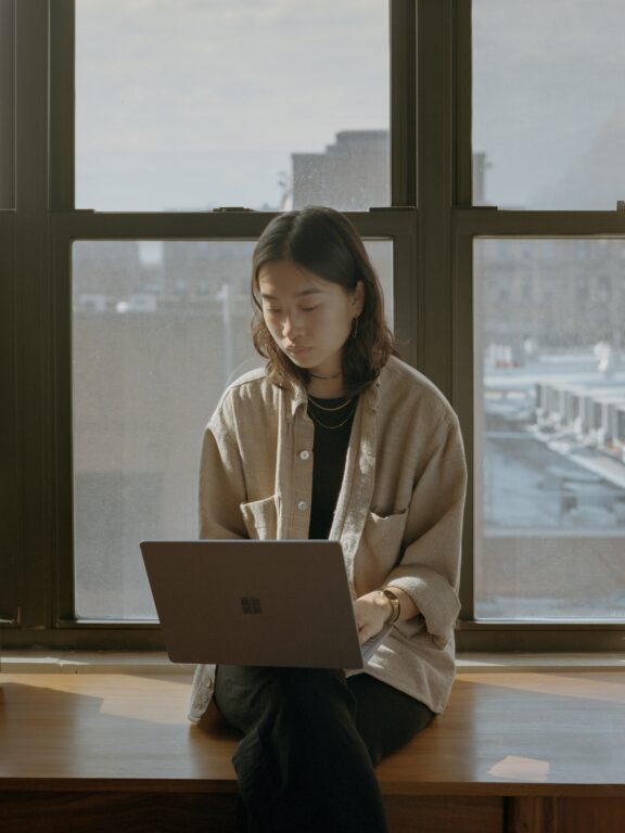 A person sits on a windowsill, looking at a laptop. City buildings are visible through large windows behind them.