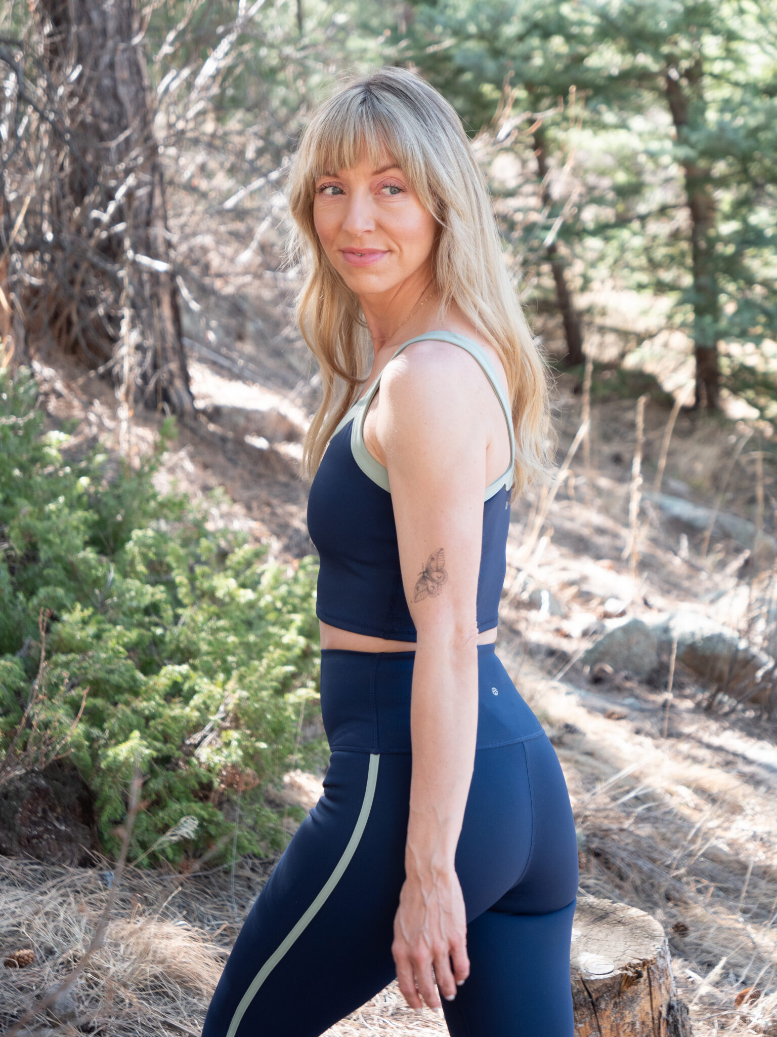 Woman with long blonde hair wearing a navy blue athletic outfit stands outdoors on a forest trail, looking over her shoulder with a slight smile.